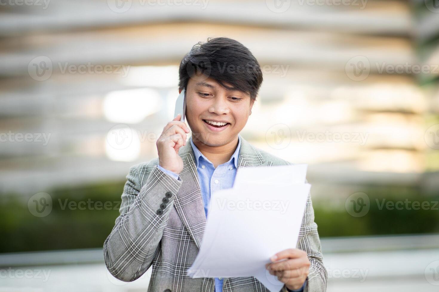 Asian man in a grey blazer is talking on a mobile phone while reading a document. He is standing outside, with a blurred background of a building and greenery photo