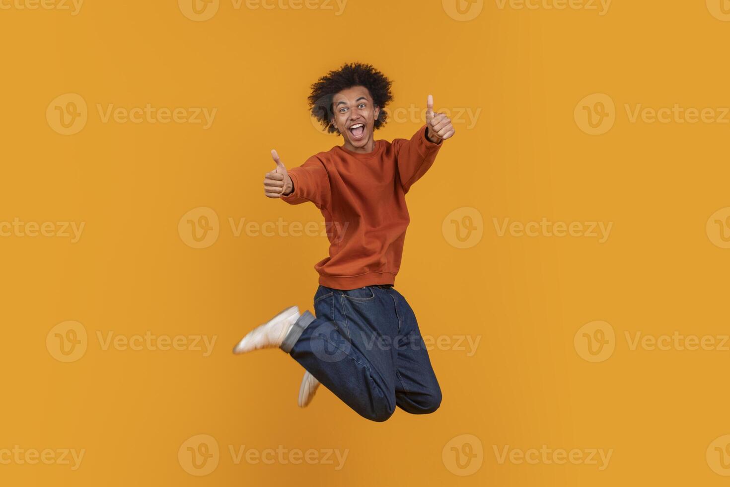 African American young man with an afro is jumping in the air with both thumbs up. He is wearing a red sweater and blue jeans. He is smiling and looking directly at the camera photo