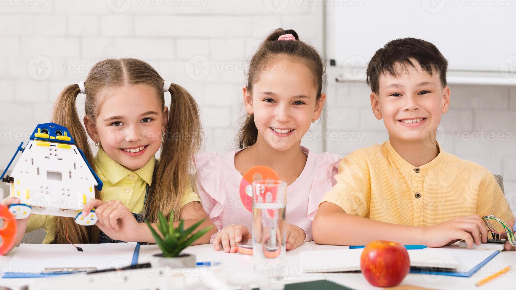 Three children, a girl holding a robot and two others, are sitting at a table in a classroom setting, smiling and working on a STEM project. photo
