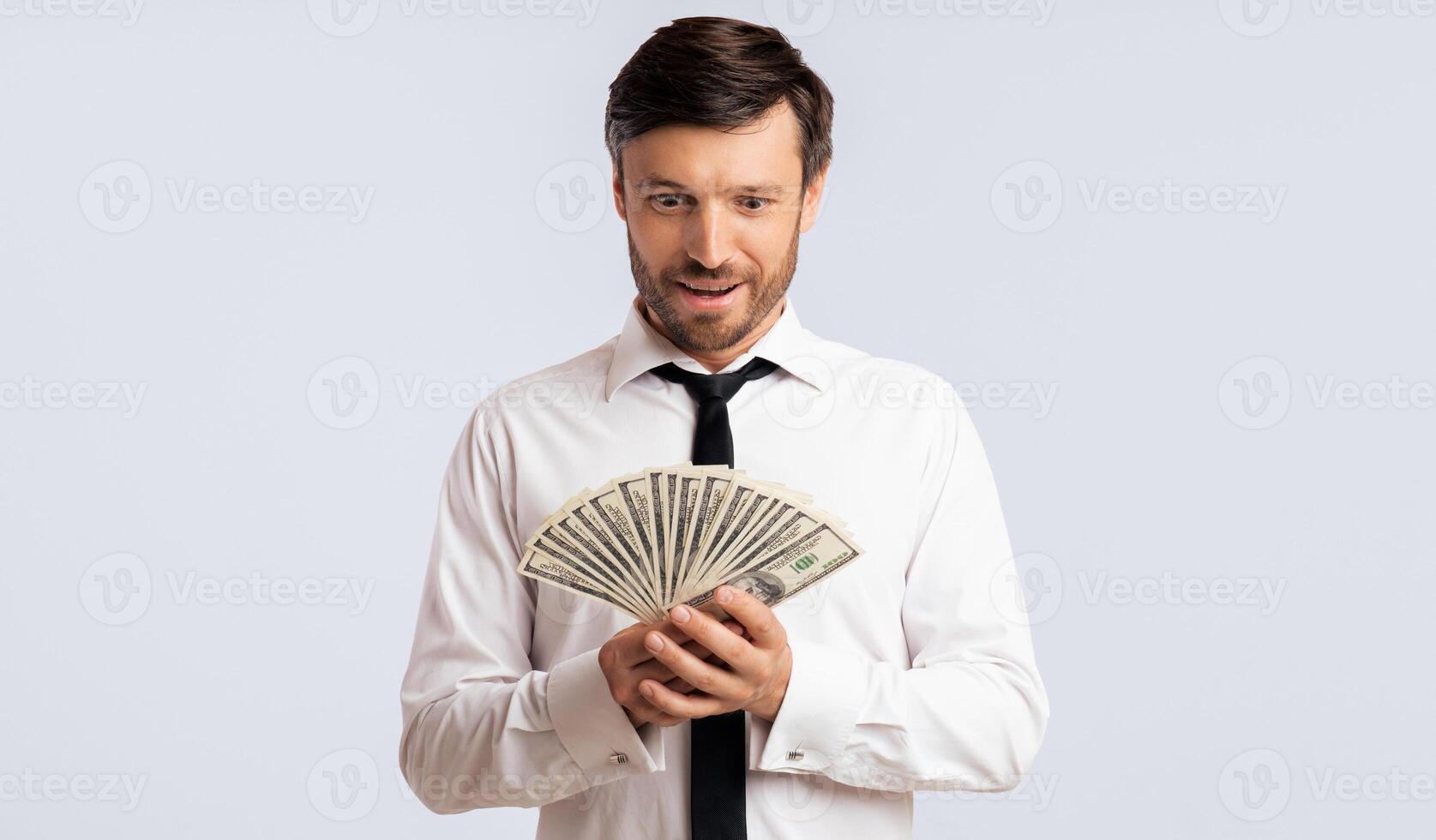 A man in a white shirt and black tie holds a fan of cash in front of him. He looks down at the money with an expression of excitement and surprise. photo