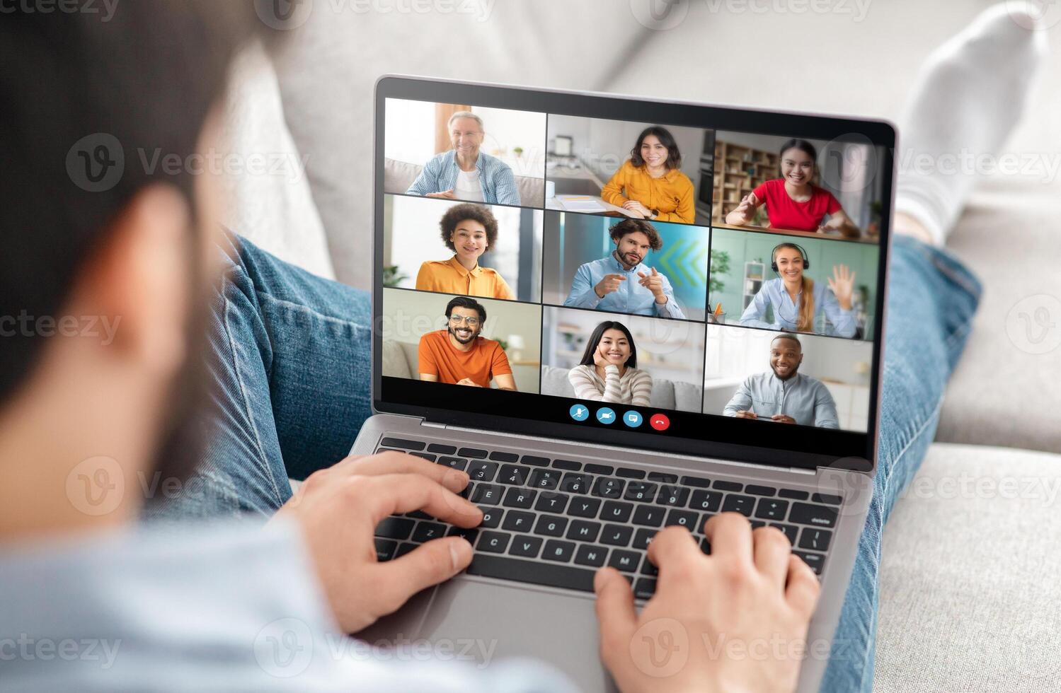 A man is sitting on a couch at home, using a laptop to participate in a conference call with several other people. He is typing on the keyboard and appears to be actively engaged photo