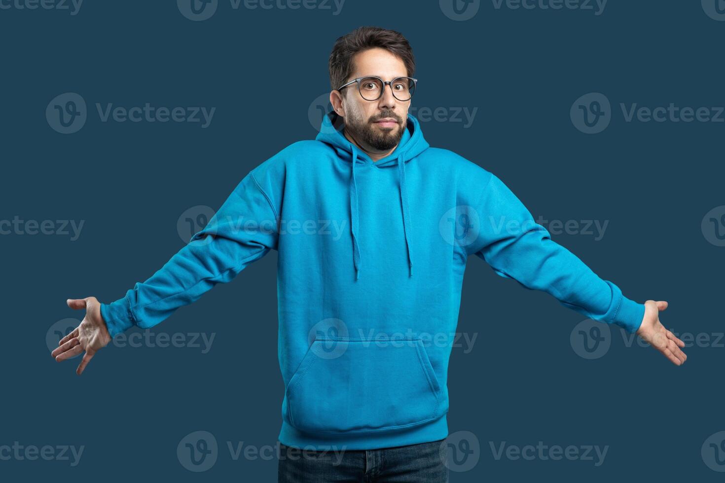 A young man stands with outstretched arms, indicating confusion or disbelief while wearing a bright blue hoodie. The background color drawing attention to his expression and attire. photo