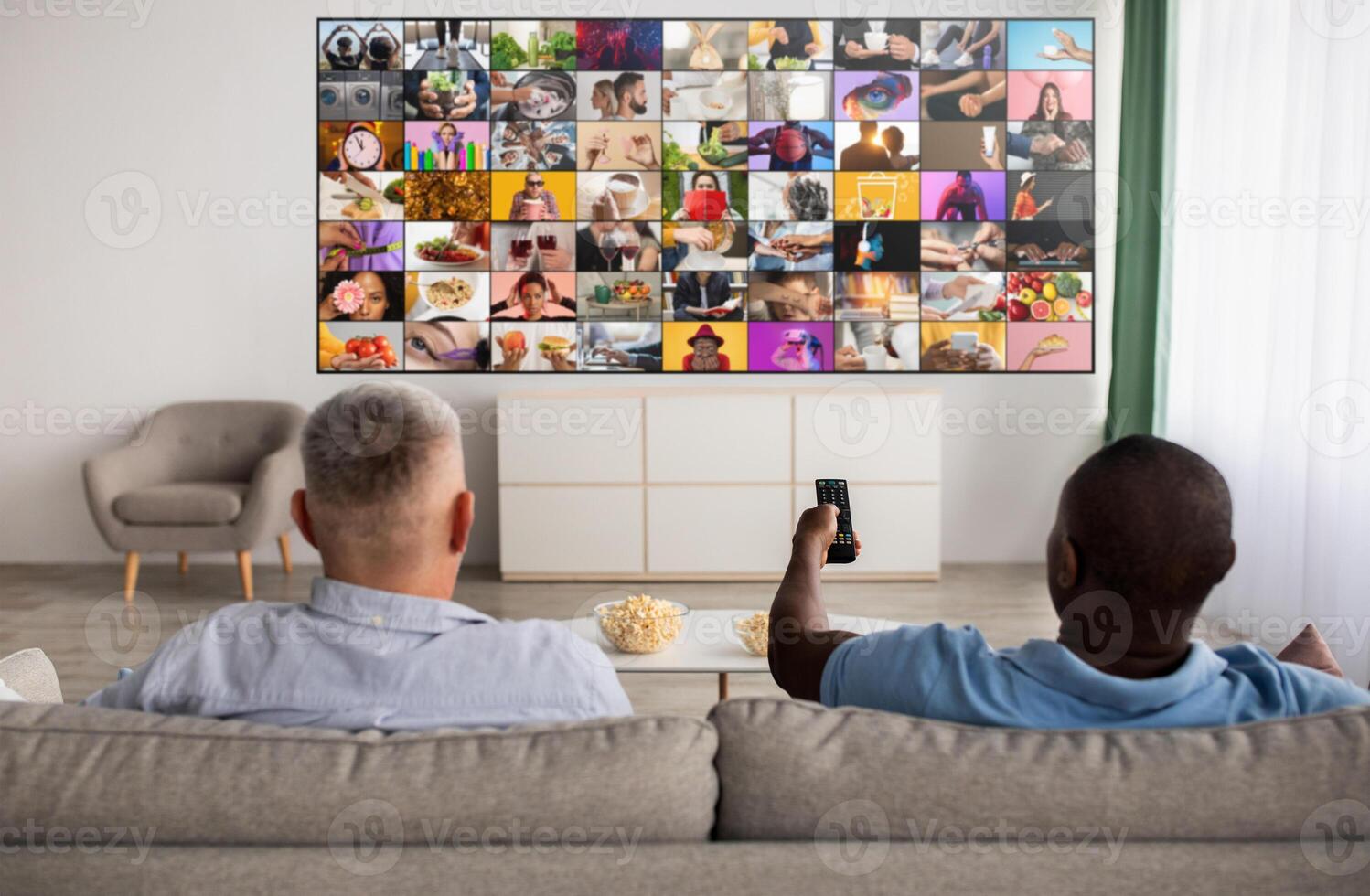 Two men are relaxing on a couch in a living room. They are watching television. The television screen is displaying a collage of many different images. One man is holding a remote control. photo