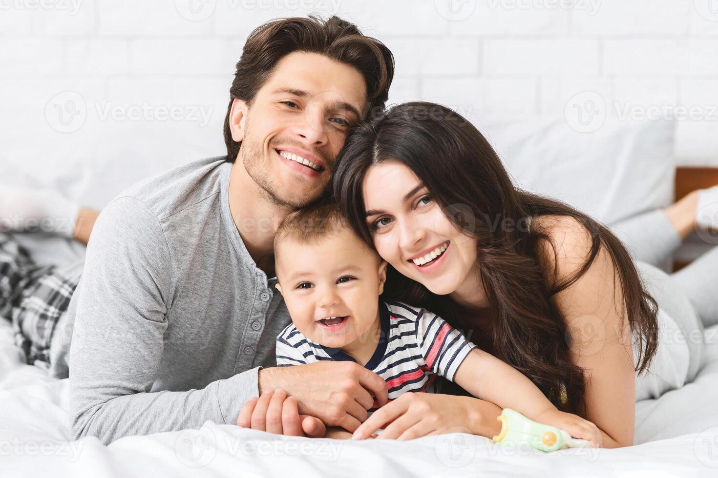 A smiling family of three lies on a bed together. The parents are both looking at the camera, while the baby is smiling at someone out of frame. The family looks relaxed and happy. photo