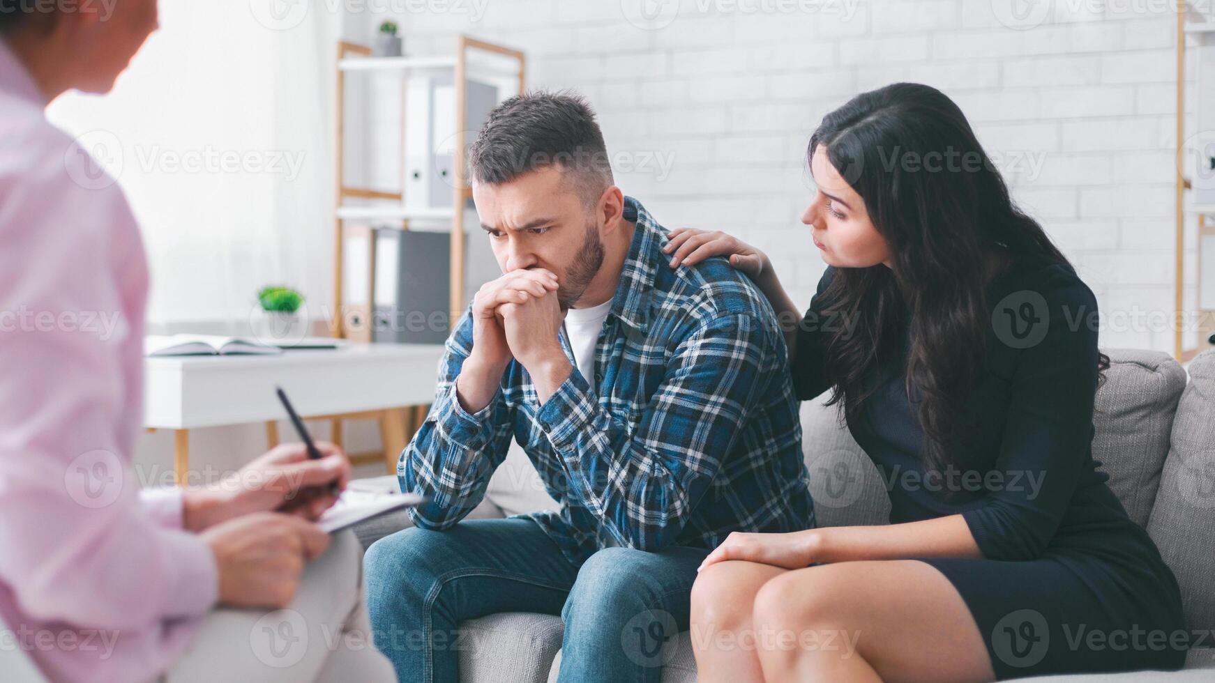 A man sits on a couch in a therapy session, looking upset and holding his hands together, while his partner sits next to him, placing a comforting hand on his shoulder photo