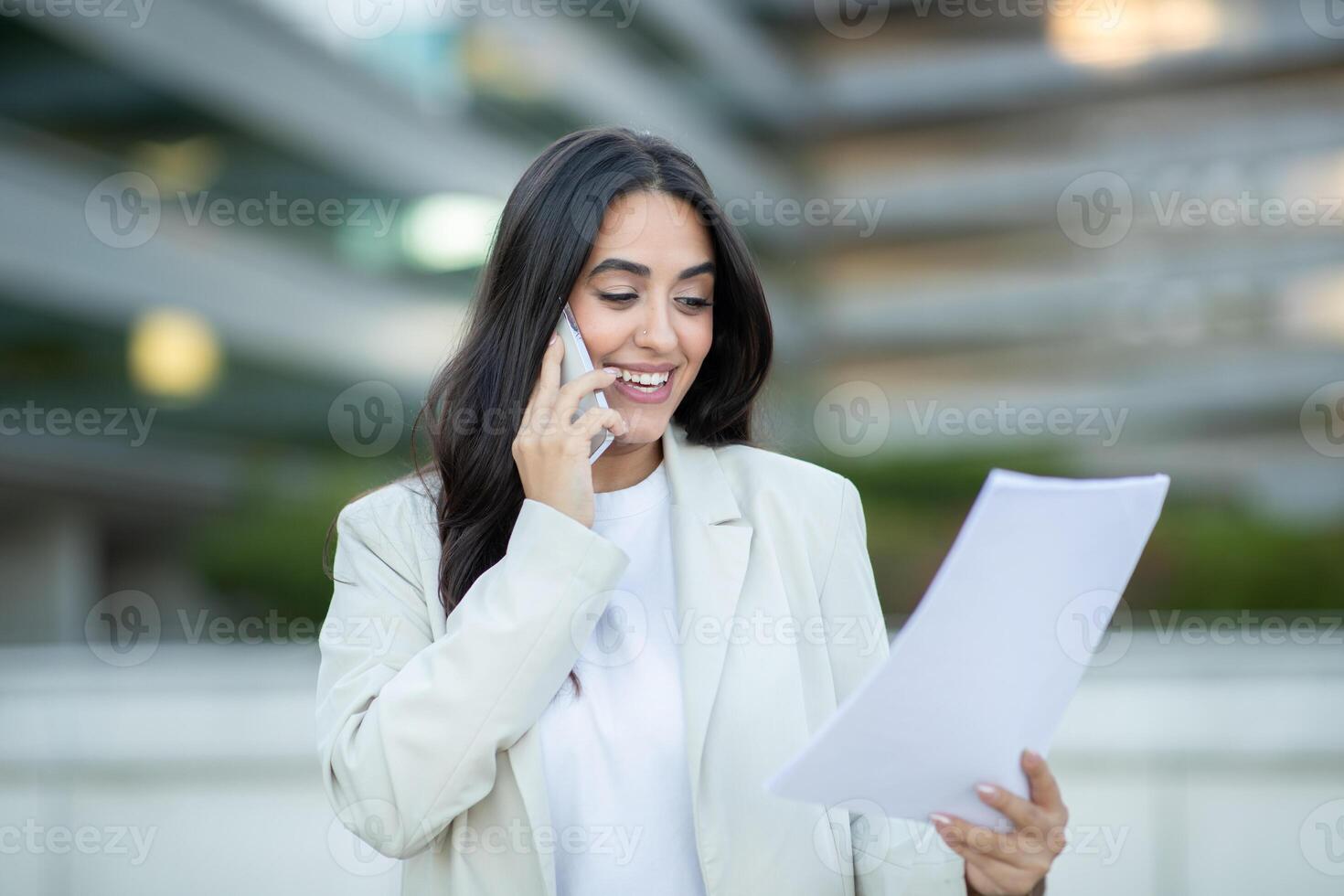 A woman in a white blazer and white top is standing outdoors, holding a document in her left hand while talking on the phone with her right hand. She is smiling and looking at the document. photo