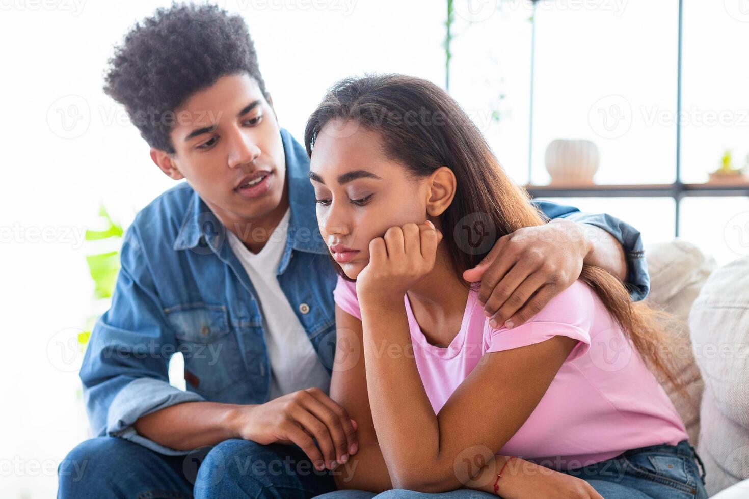 A teen guy sits next to a girl who is looking down with a sad expression. He has his arm around her shoulder as if offering comfort. The image suggests a moment of emotional support photo