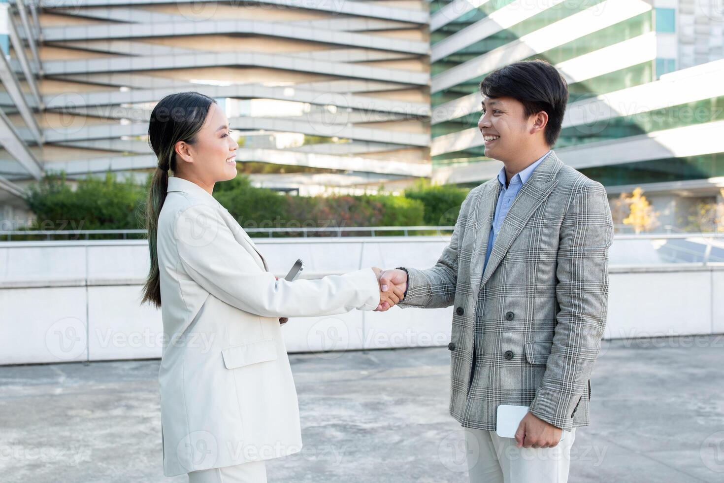 Two Asian business professionals man and woman are having a friendly handshake outside a contemporary high-rise office building. The urban environment reflects a dynamic workplace setting. photo