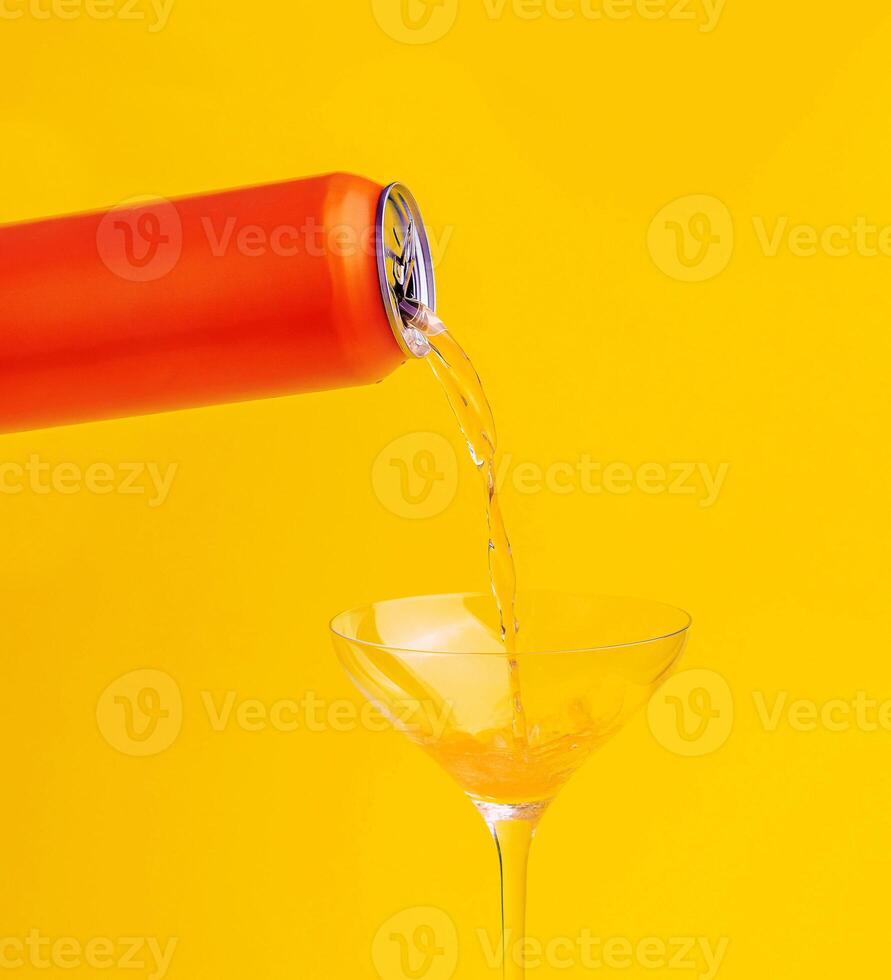 Pouring orange drink from aluminum can into cocktail glass on yellow background photo