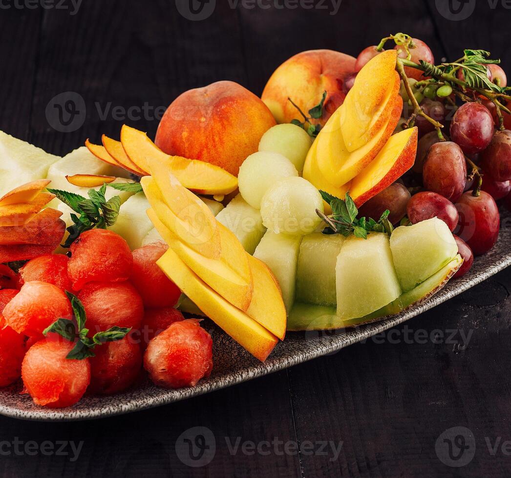 Oval plate overflowing with fresh watermelon, cantaloupe, peaches and grapes on dark wooden table photo