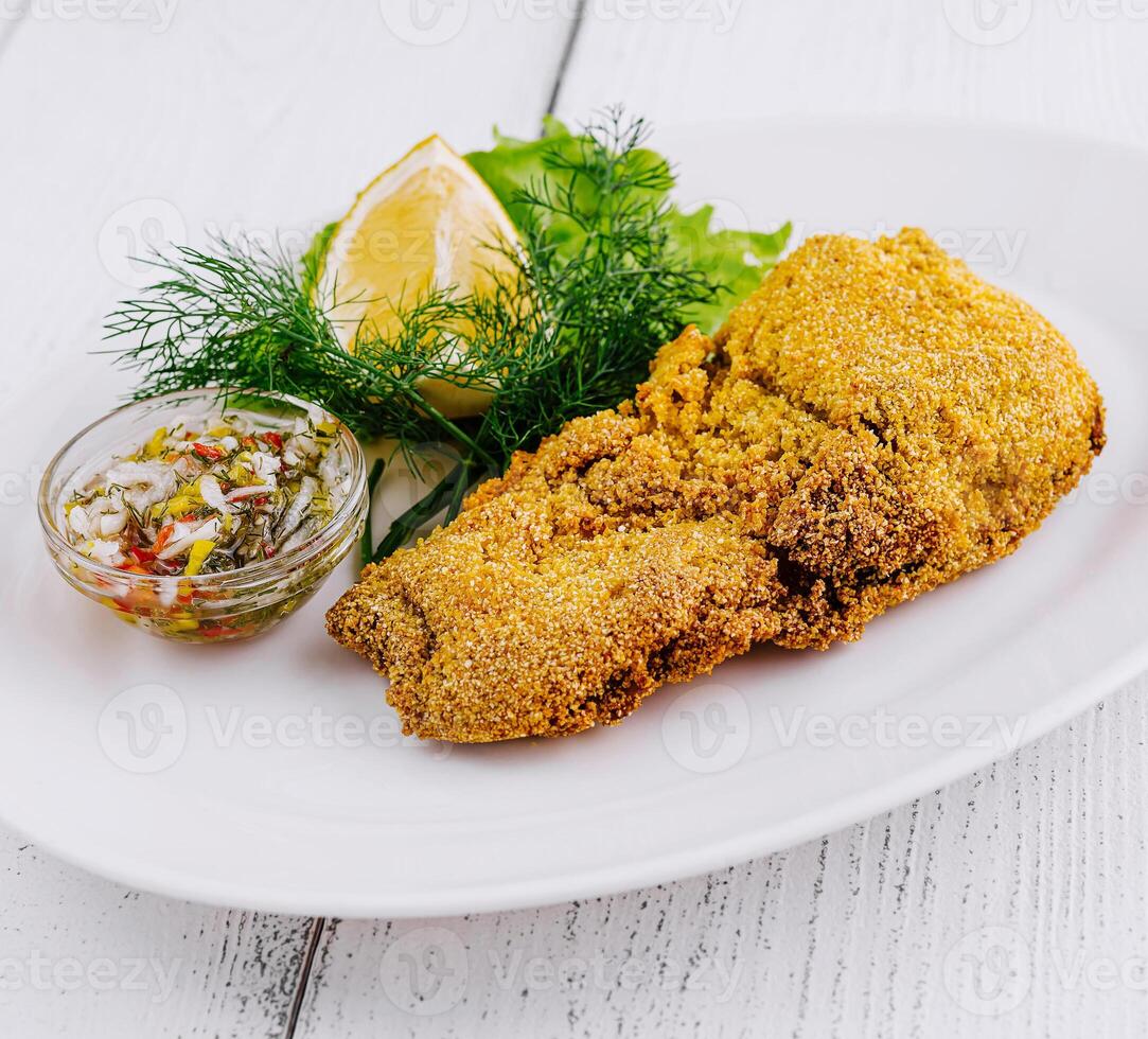 Breaded fish fillet being served on white plate with lemon and greens photo