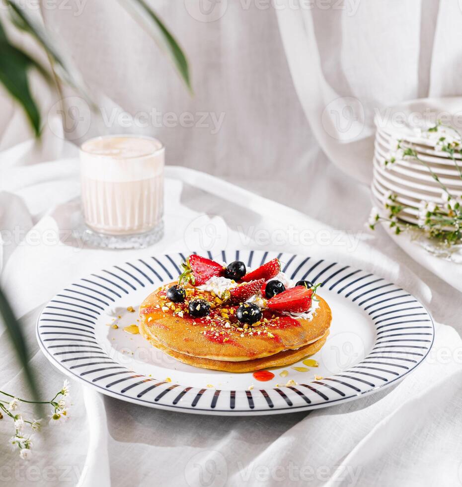 Stack of delicious pancakes with whipped cream, berries and pistachios is served on white tablecloth photo