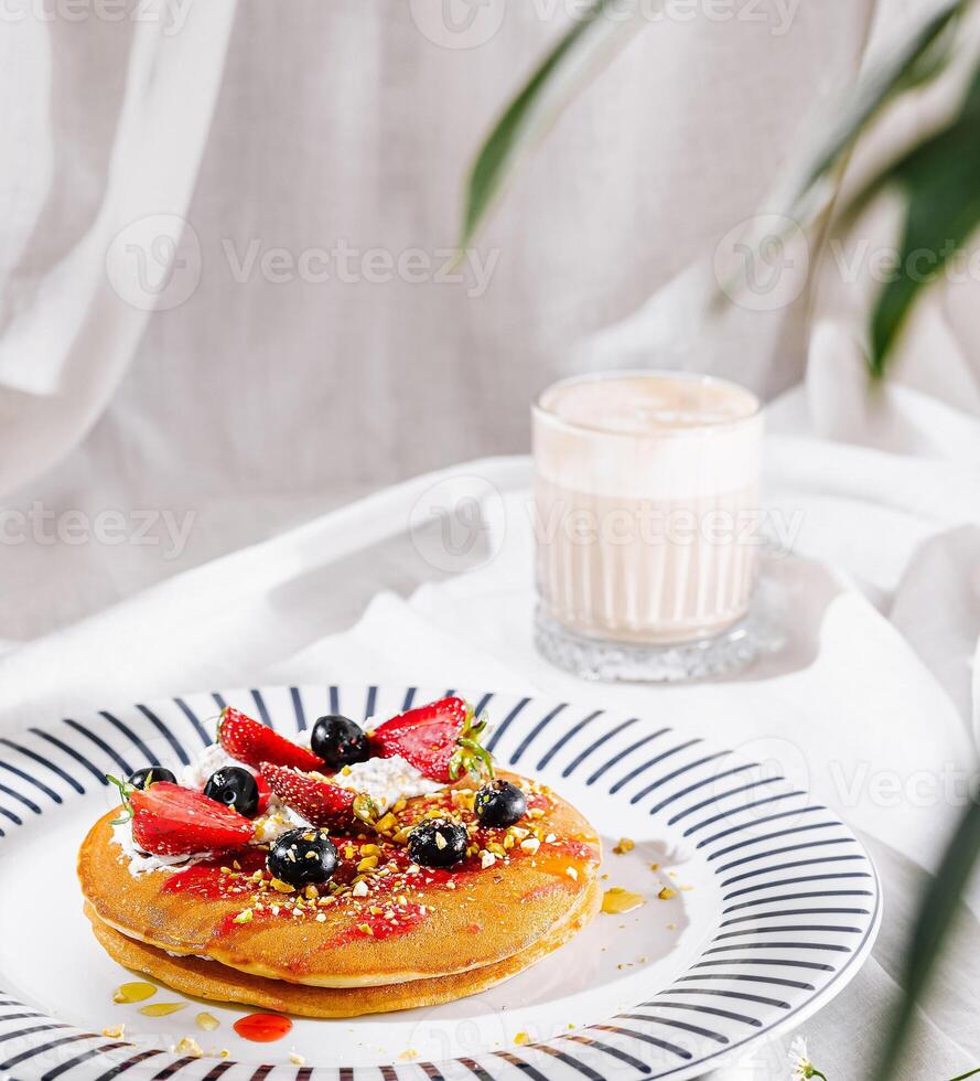Stack of delicious pancakes with whipped cream, berries and pistachios is served on white tablecloth photo