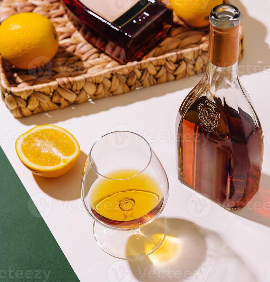 Bottle and glass of cognac with lemons on white and green background photo