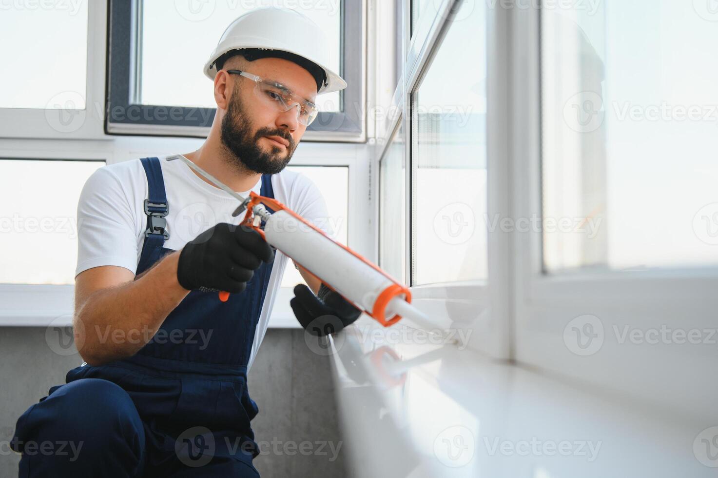Young man, dressed in a uniform, doing repair in the house. Handyman is applying silicone from a caulk gun to prevent water leak photo