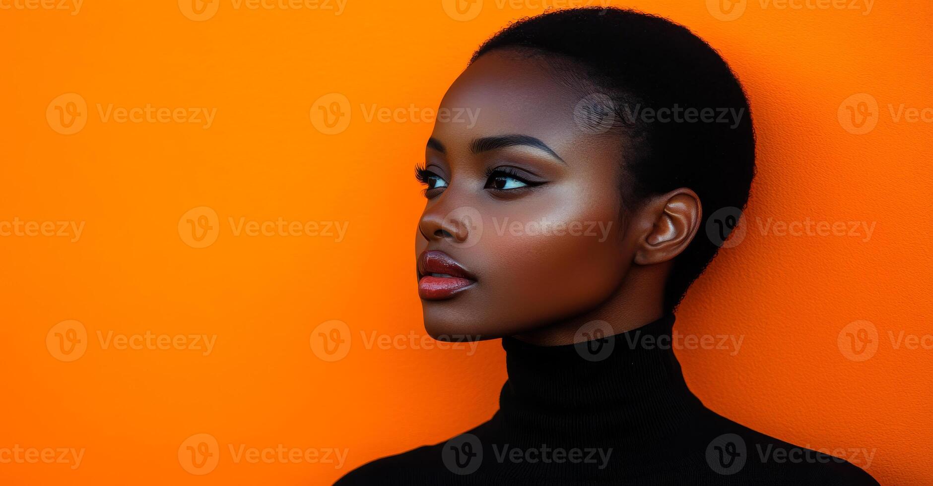 A woman with a short haircut and a black sweater stands in front of an orange wall. She has a serious expression on her face photo
