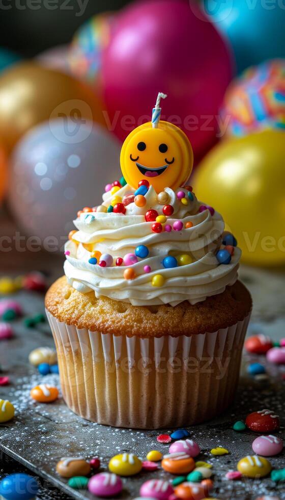 A cupcake with a smiley face on top and a candle in the middle. The cupcake is decorated with colorful sprinkles and is placed on a table with a bunch of balloons. The image conveys a festive photo