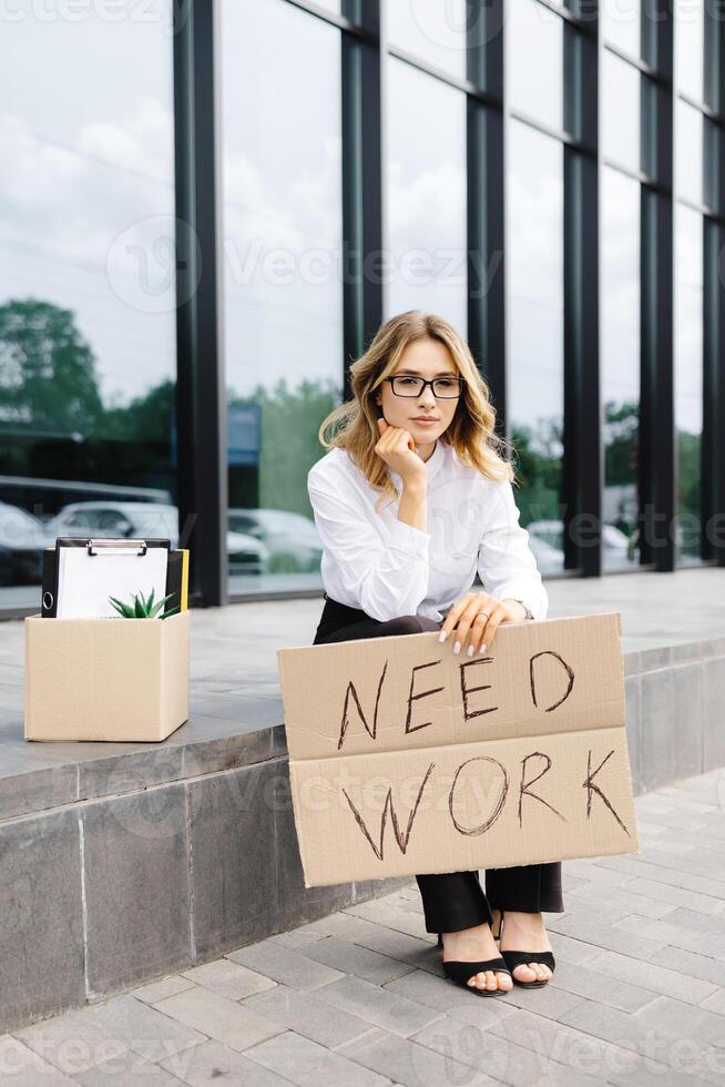 Woman sitting outdoor with cardboard banner with the slogan Need work next to box of stuff. Beautiful female protester at strike against unemployment photo