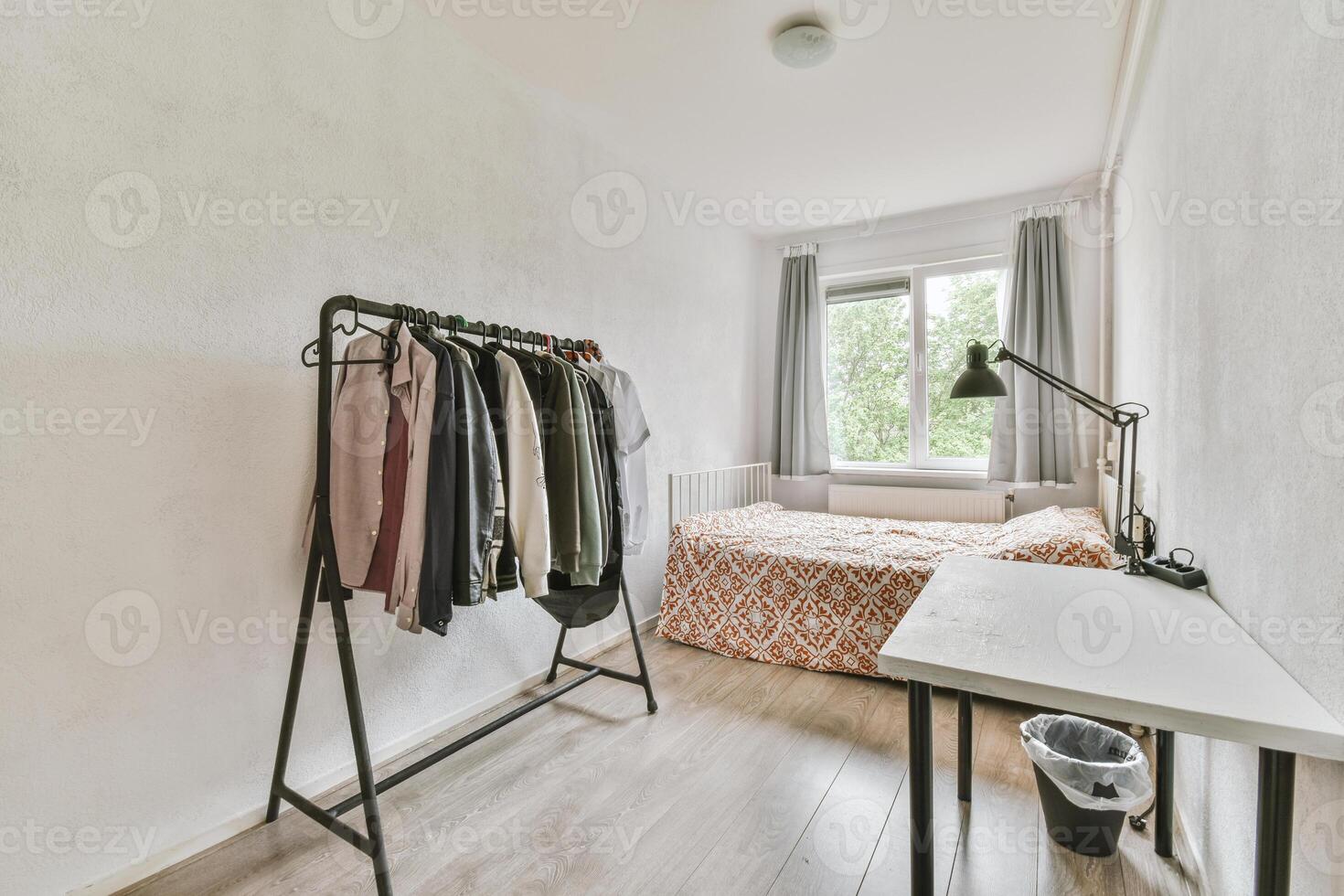 A bedroom with a bed, clothes rack and a desk photo