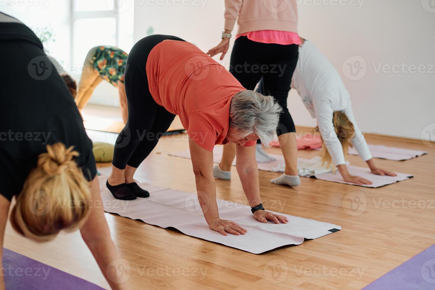 A group of senior women engage in various yoga exercises, including neck, back, and leg stretches, under the guidance of a trainer in a sunlit space, promoting well-being and harmony photo