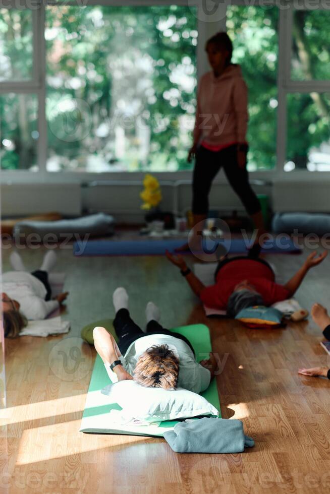 A group of senior women engage in various yoga exercises, including neck, back, and leg stretches, under the guidance of a trainer in a sunlit space, promoting well-being and harmony photo