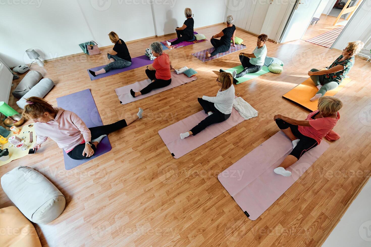 A group of senior women engage in various yoga exercises, including neck, back, and leg stretches, under the guidance of a trainer in a sunlit space, promoting well-being and harmony photo