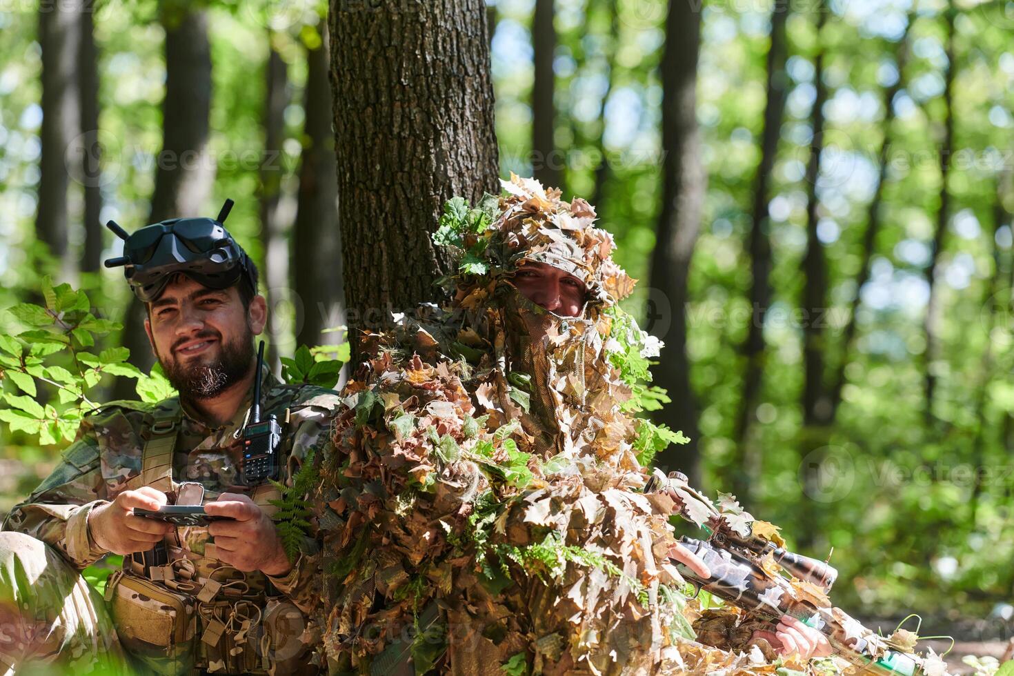 A skilled sniper and a soldier operating a drone with VR goggles strategize and observe the military action while concealed in the forest photo
