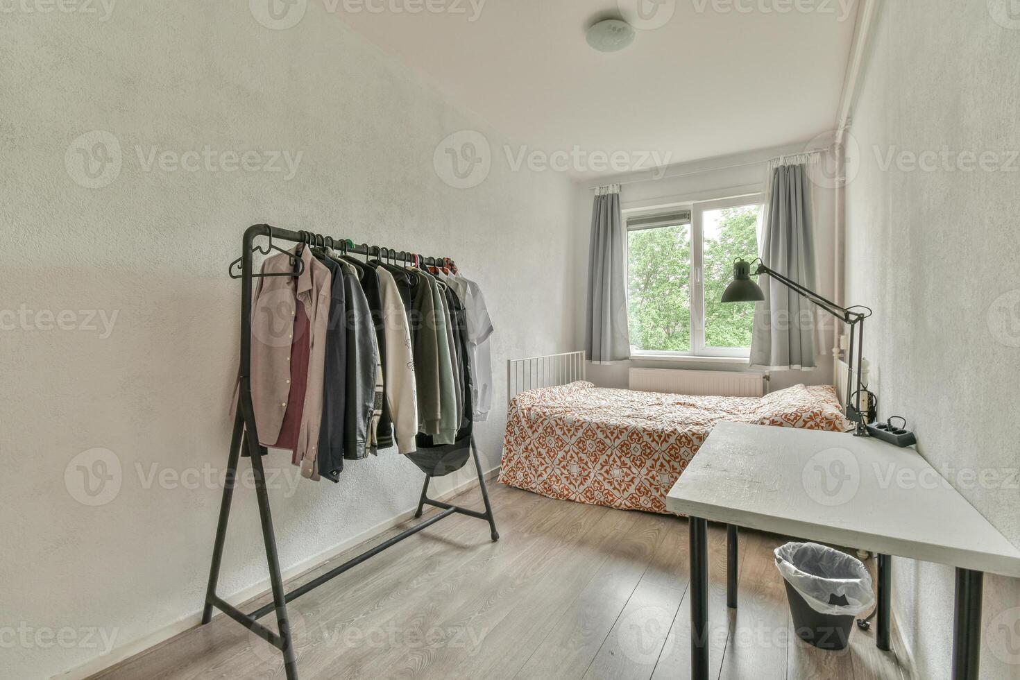 A bedroom with a bed, clothes rack and a desk photo