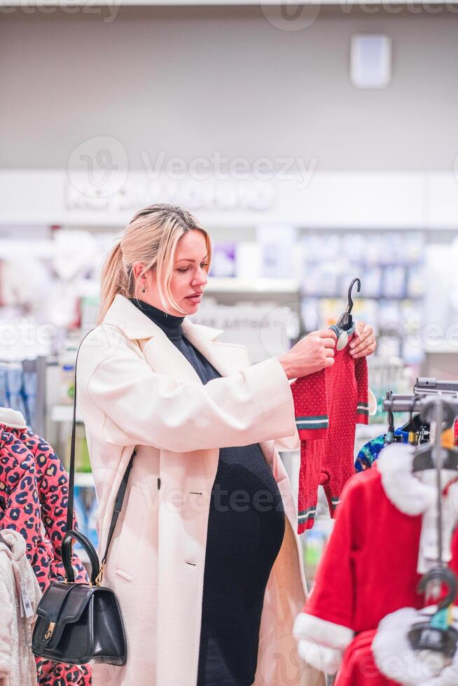 Young pregnant woman choosing newborn clothes at baby shop store photo