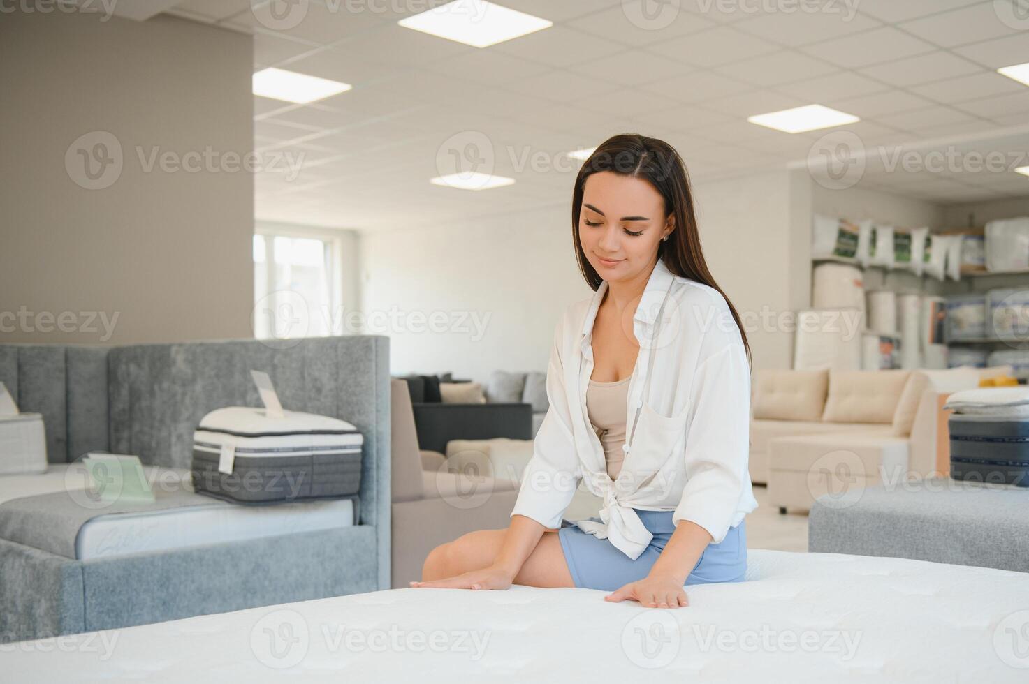 happy brunette female is sitting on mattress for testing it in the store photo