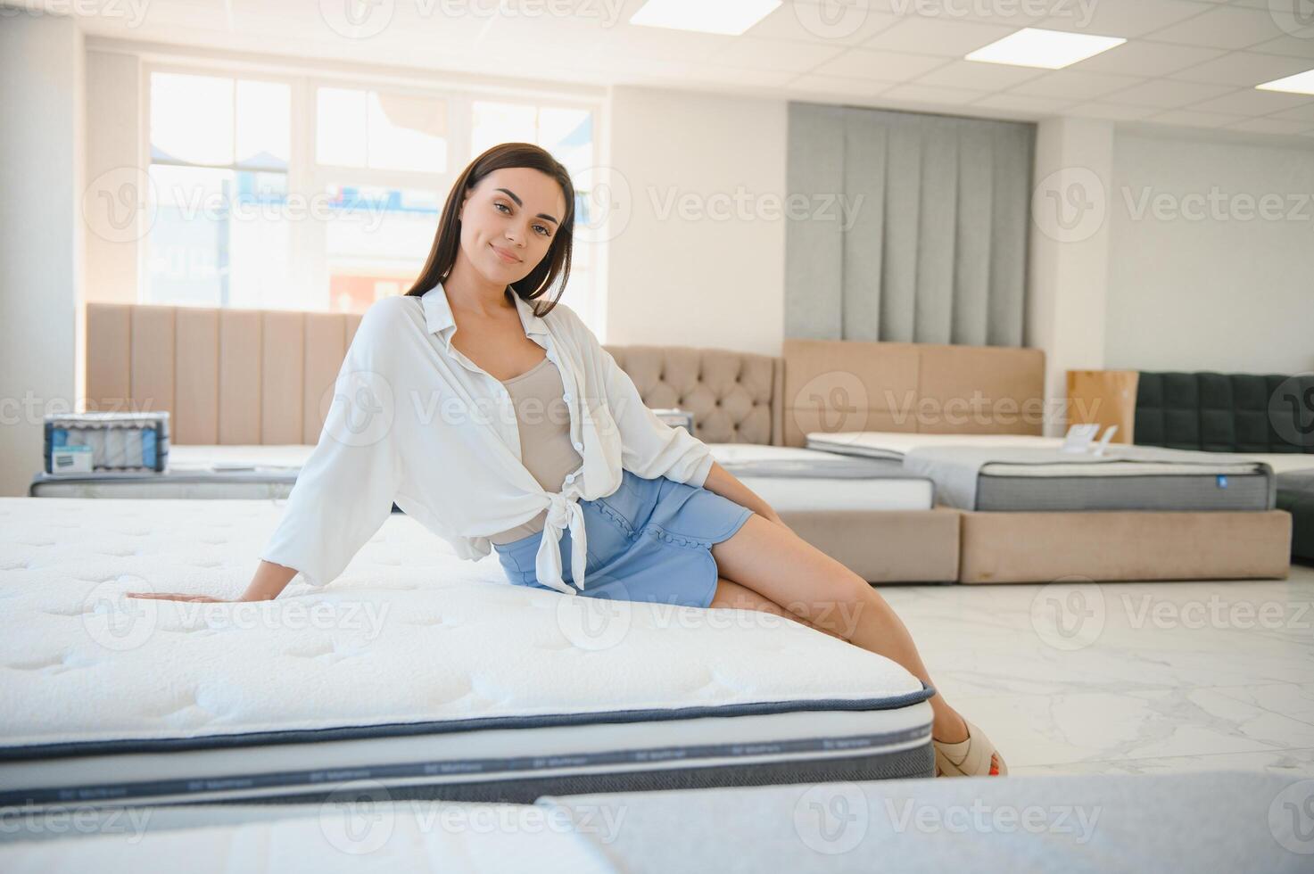 happy brunette female is sitting on mattress for testing it in the store photo