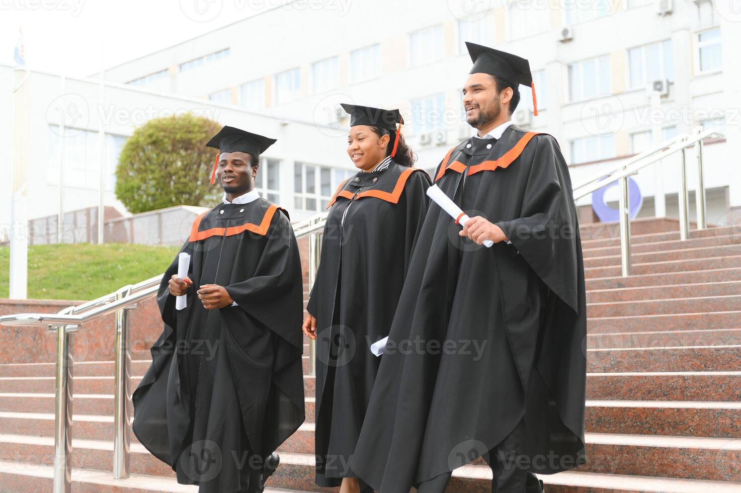 Group of happy smiling multicultural people in graduation gowns and caps with diplomas in hands graduate university outdoors in campus looking at camera. Graduation from college, university concept. photo
