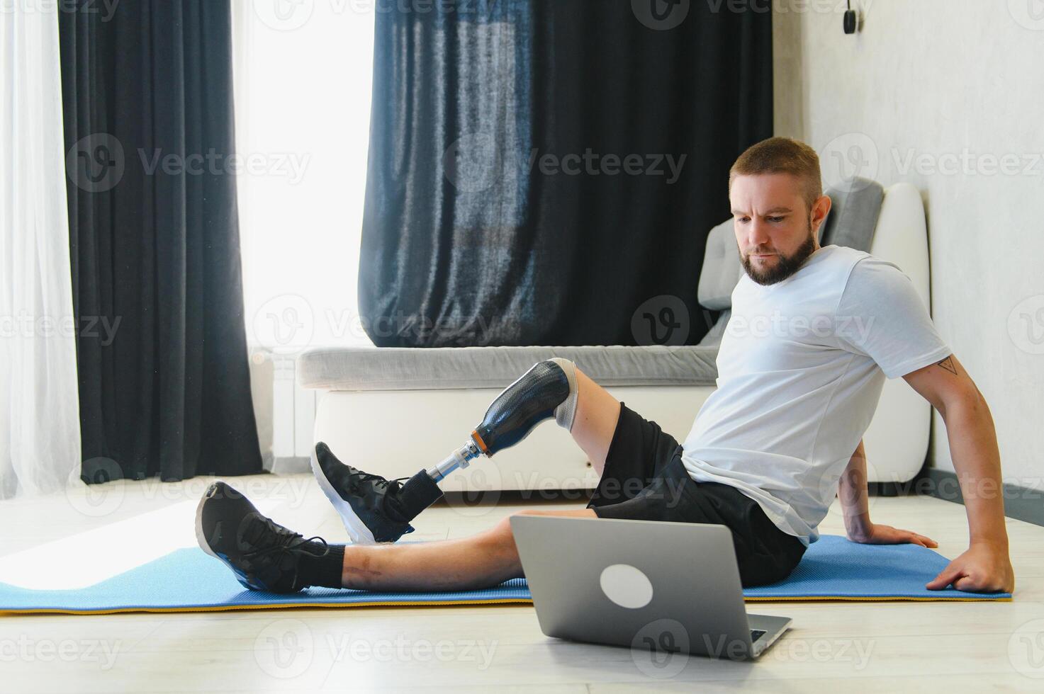 Sporty fit young man amputee with prosthetic leg disability prosthesis doing training exercises on mat at home using laptop. Inclusive online sport classes for people with disabilities concept photo