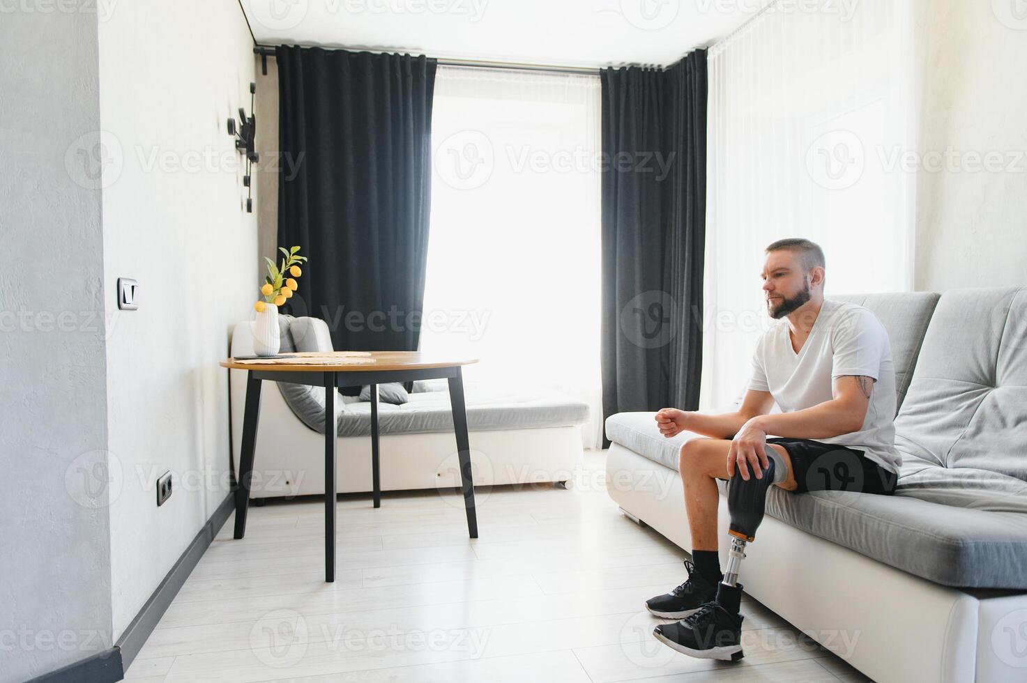 Thoughtful young pensive man amputee with prosthetic leg disability above knee transfemoral leg prosthesis sitting on sofa at home. People with amputation disabilities everyday life photo