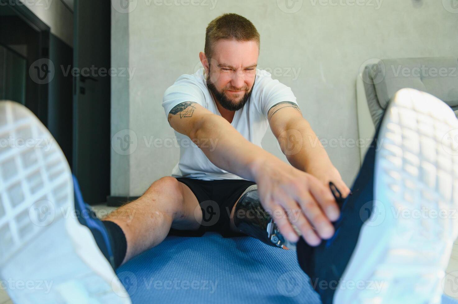 Man amputee with prosthetic leg disability on transfemoral leg prosthesis artificial device making stretching rehabilitation physical exercise at home. People w amputation disabilities everyday life photo