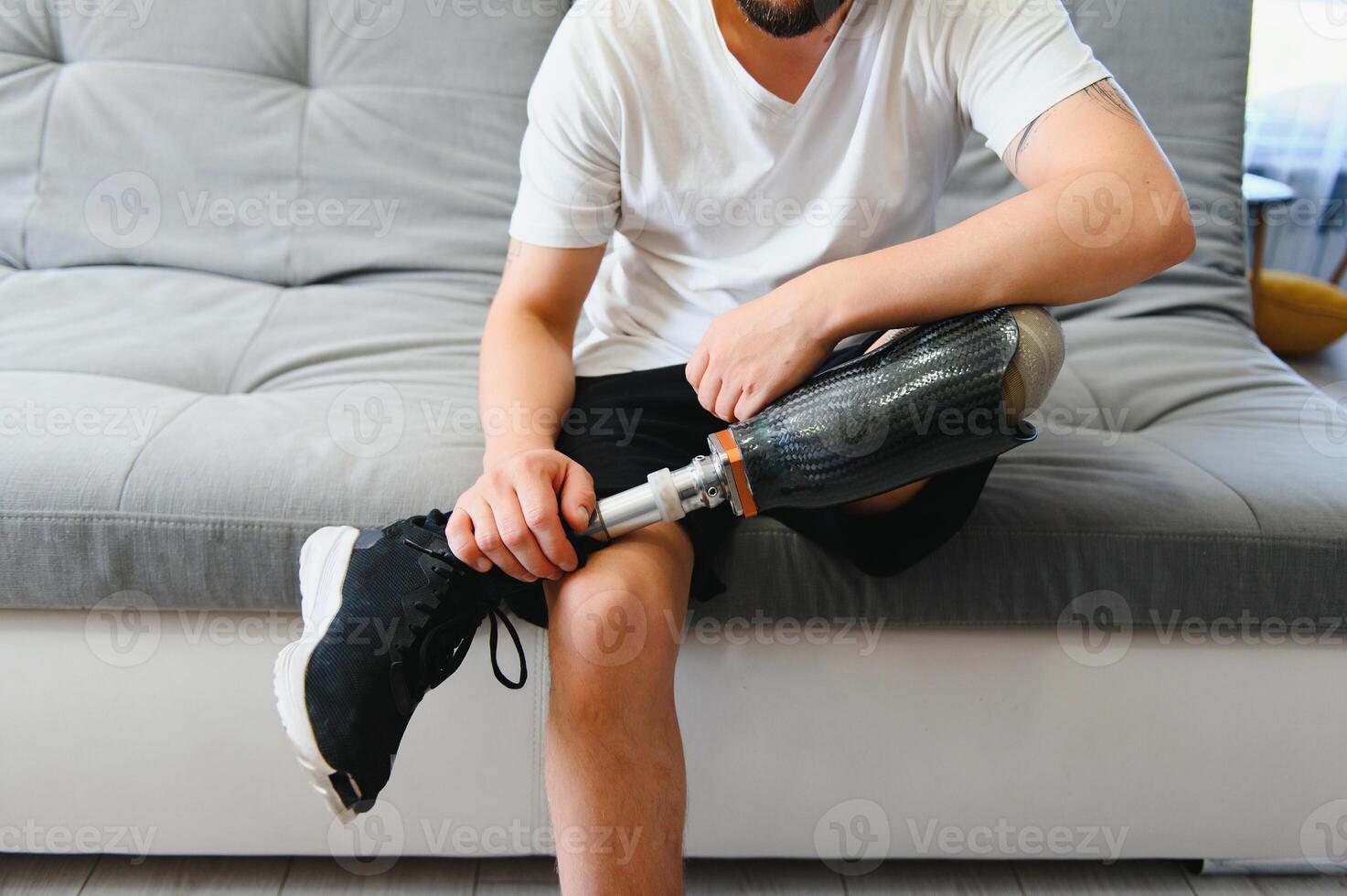 Man amputee with prosthetic leg disability on above knee transfemoral leg prosthesis wearing shoe sitting on sofa, close up. People with amputation disabilities everyday life concept photo