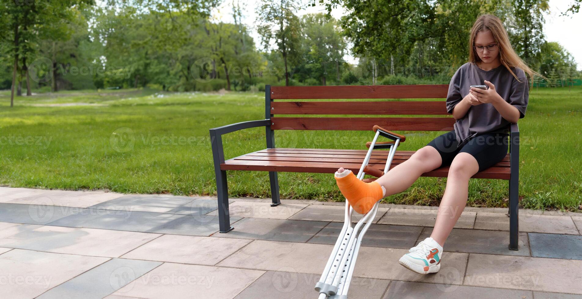 Teenage girl with a leg in a cast seated on a park bench, utilizing her smartphone to browse social media and content while surrounded by the tranquil outdoor environment photo