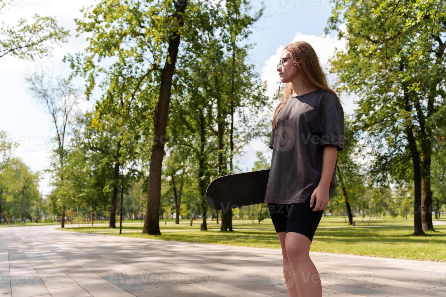 Carefree teenage skateboarder with windswept locks, wearing a casual gray tee, holding her board in a serene summer park backdrop, providing ample space for text overlay photo