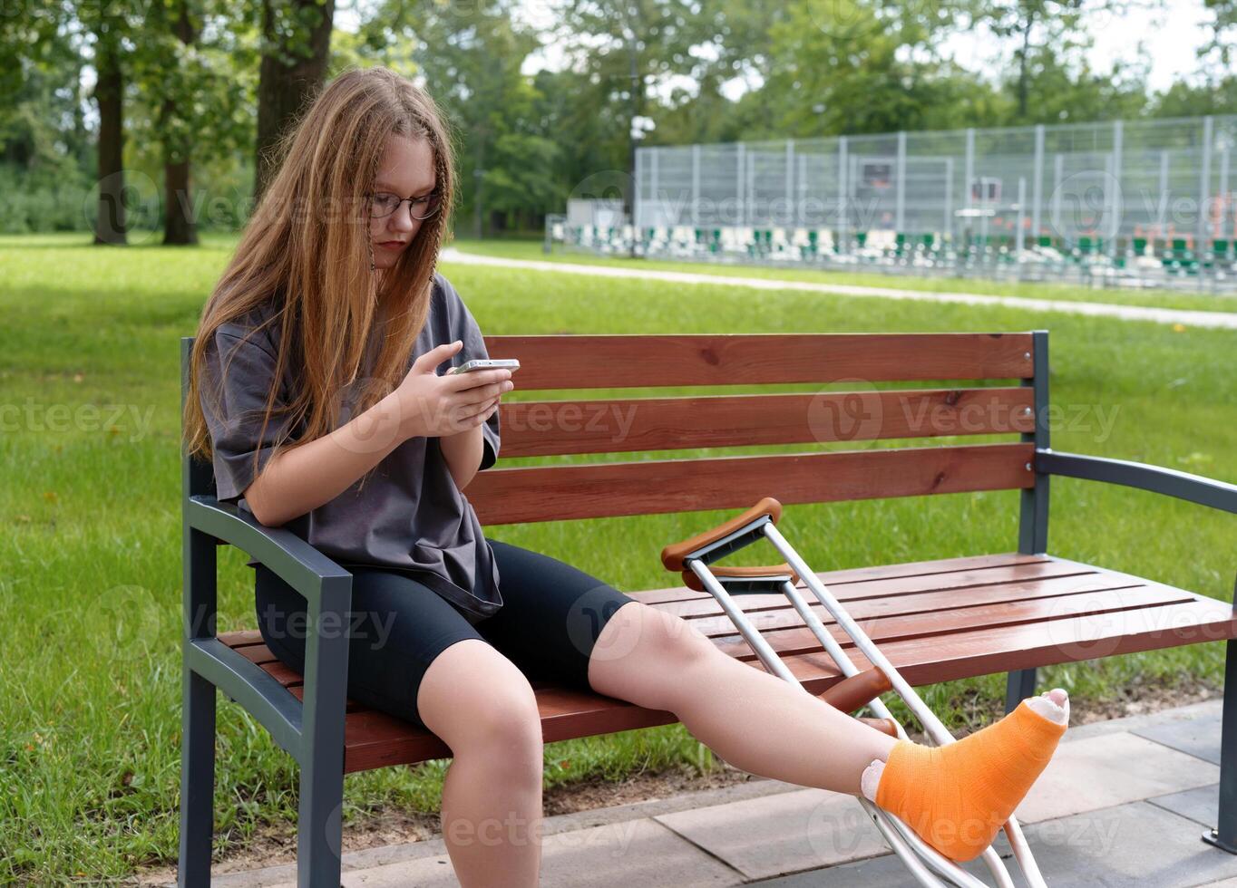 Teenage girl with a leg cast sitting on a park bench, using her smartphone to engage with social media as she recovers in the summer setting photo