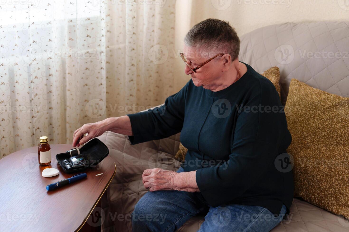 portrait captures an older adult with glasses and short gray hair sitting on a sofa, focused on the task of using a glucometer to measure her blood sugar, a crucial step in her diabetes care photo