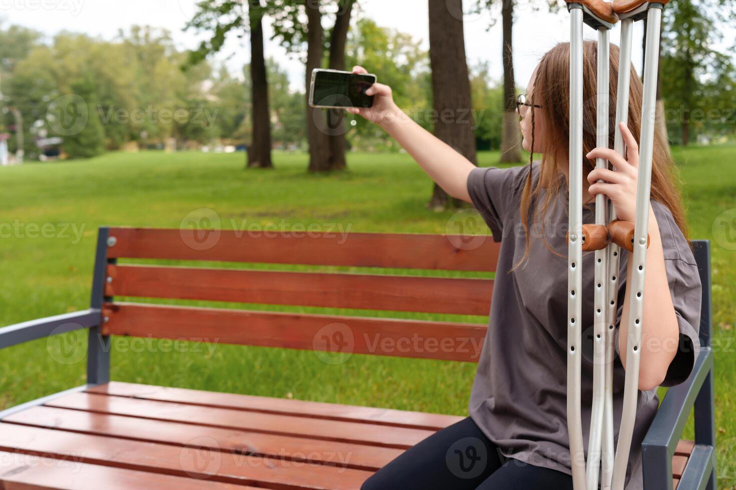 Young female with a casted leg relaxing on a bench, pausing to snap a selfie as she enjoys the peaceful outdoor environment during her rehabilitation photo