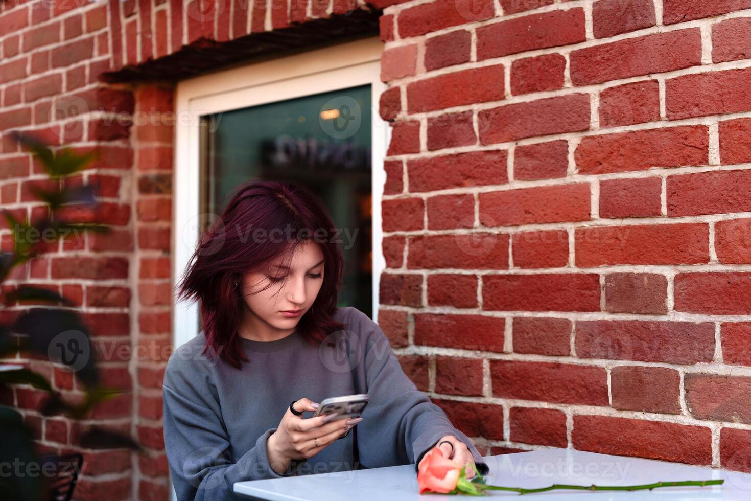 In a cozy cafe setting, a teenager with striking hair color rests her gaze on her mobile screen, a single rose on the table suggesting an introspective moment of solitude photo