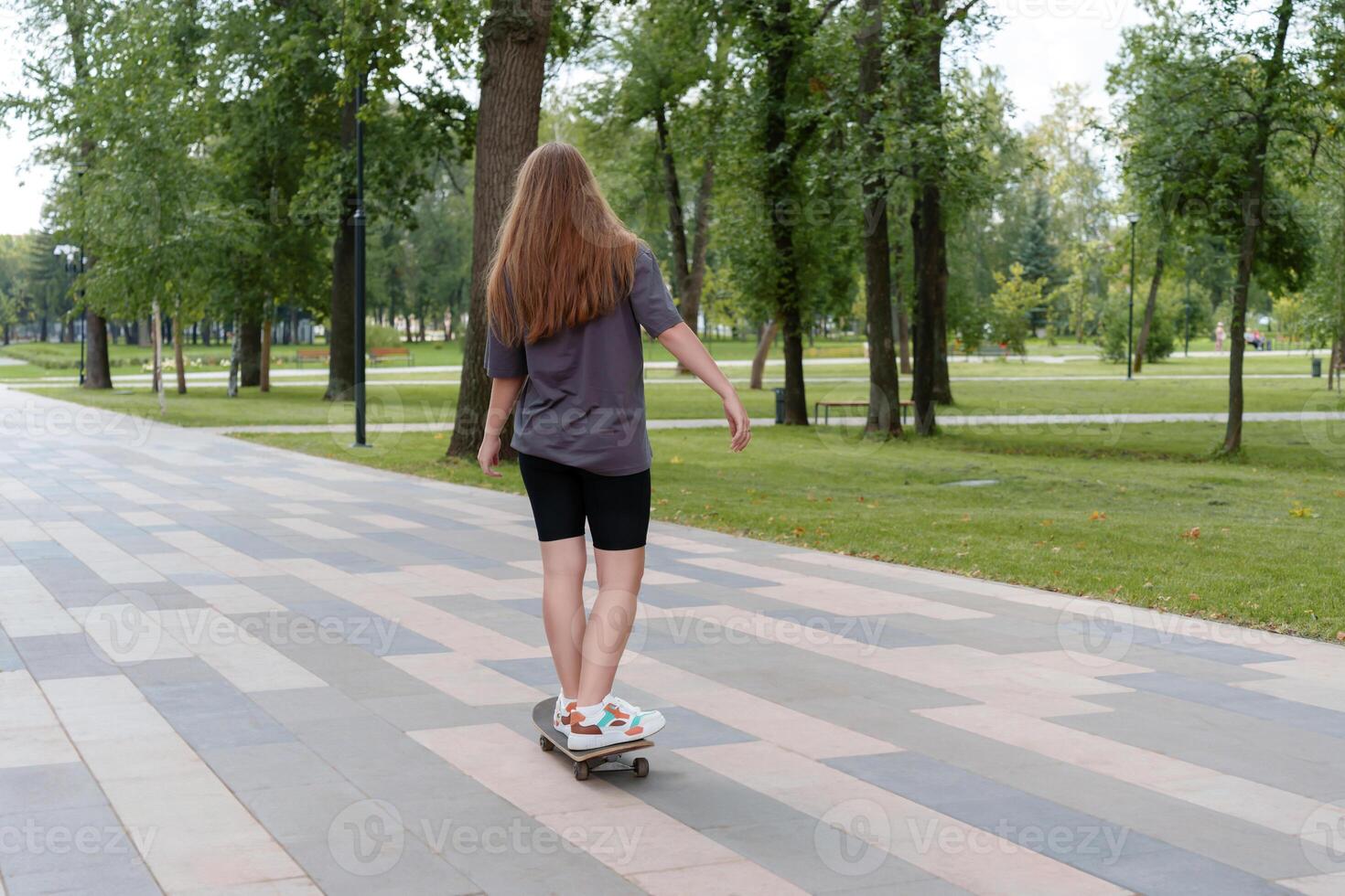 Rear view of a teenage girl with long, free-flowing hair riding a skateboard on the pavement in a green, tranquil park setting photo