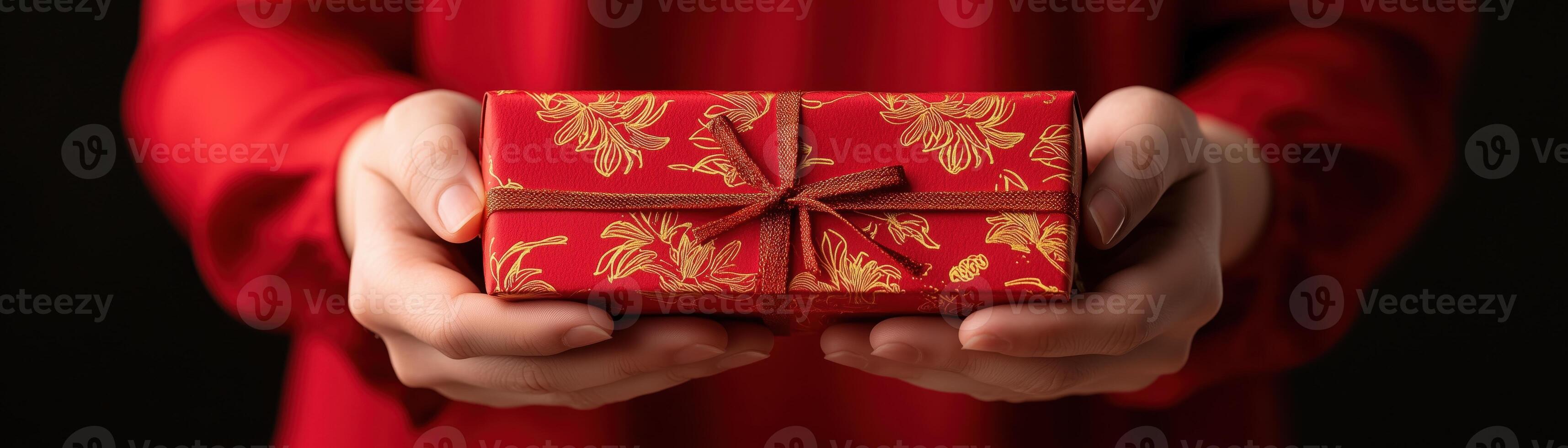 A person holding a beautifully wrapped red gift box adorned with gold patterns, symbolizing celebration and joy, against a dark background, creating a warm festive atmosphere. photo