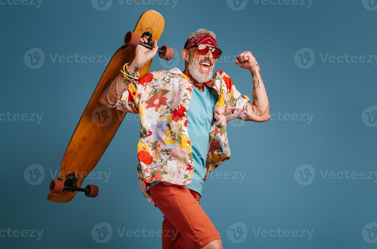 Cool senior man in funky shirt carrying longboard and smiling against blue background photo