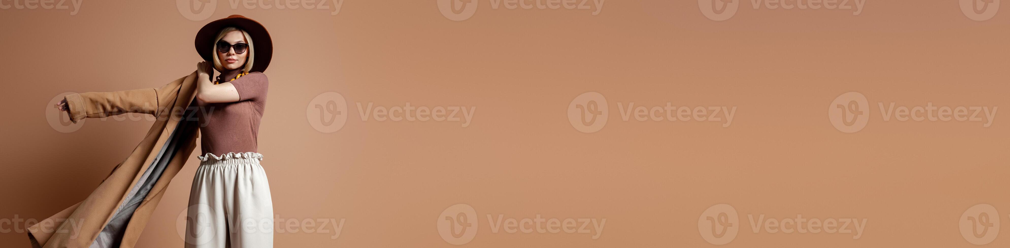 Fashionable young woman in elegant hat wearing coat against brown background photo