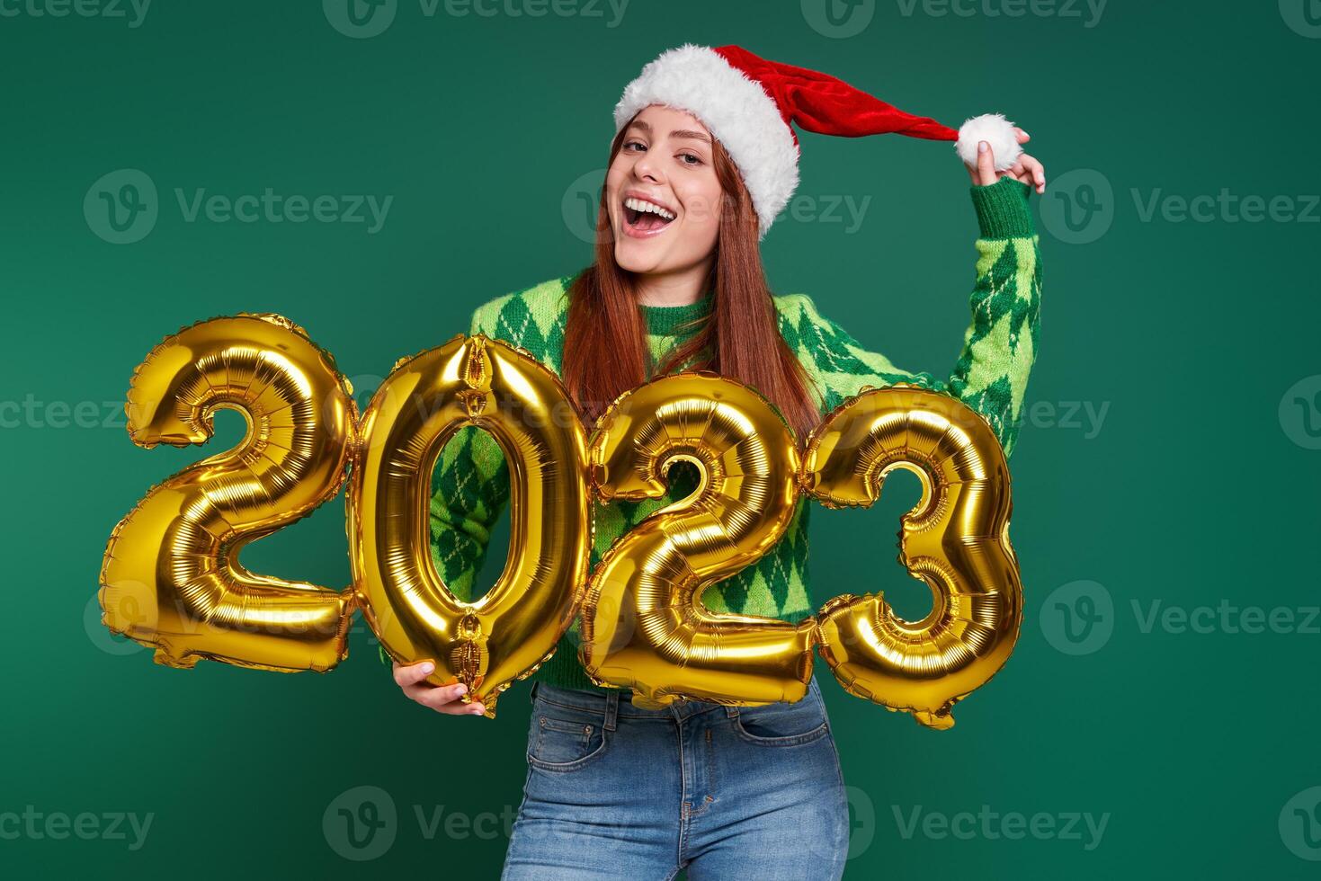 Excited young woman in Christmas hat holding gold colored numbers against green background photo