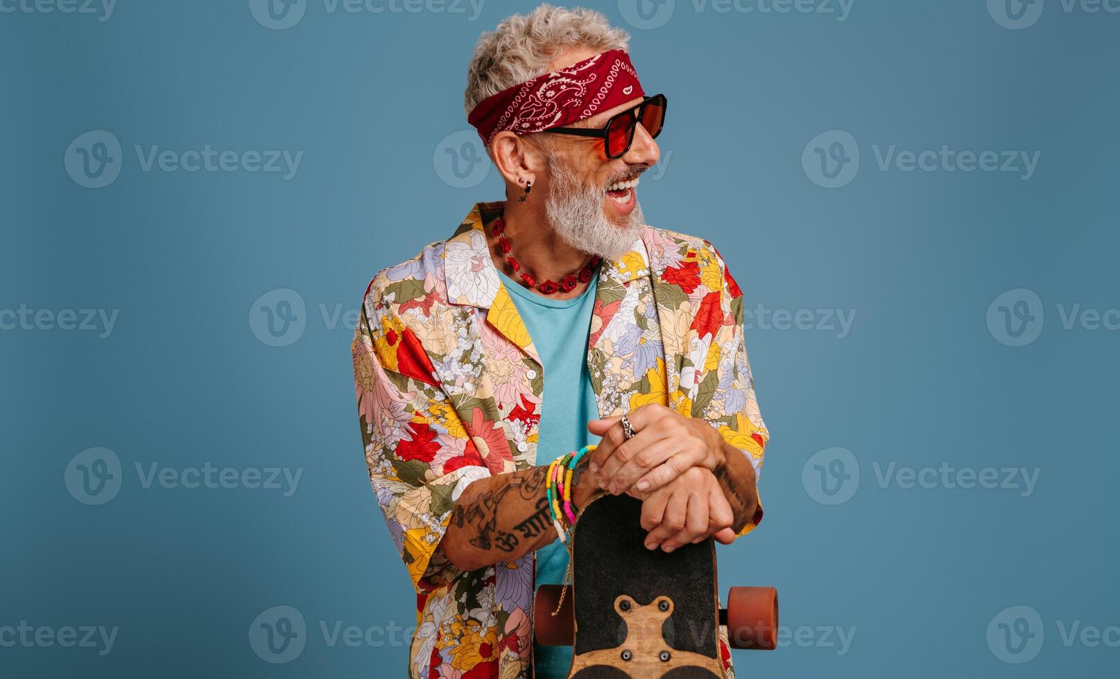 Stylish mature man in funky shirt leaning on longboard and laughing against blue background photo