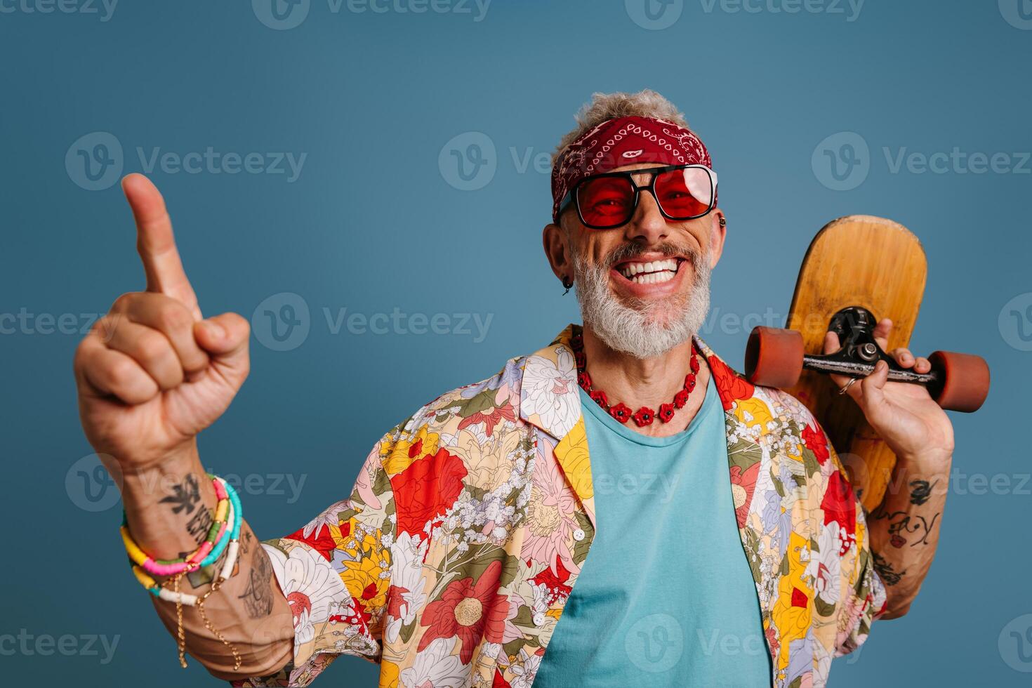 Joyful hipster mature man in stylish funky shirt carrying longboard and gesturing against blue background photo