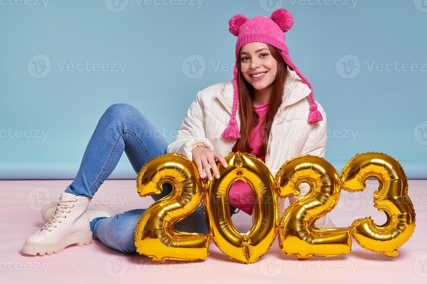 Beautiful young woman in winter coat holding gold colored numbers while sitting against blue background photo